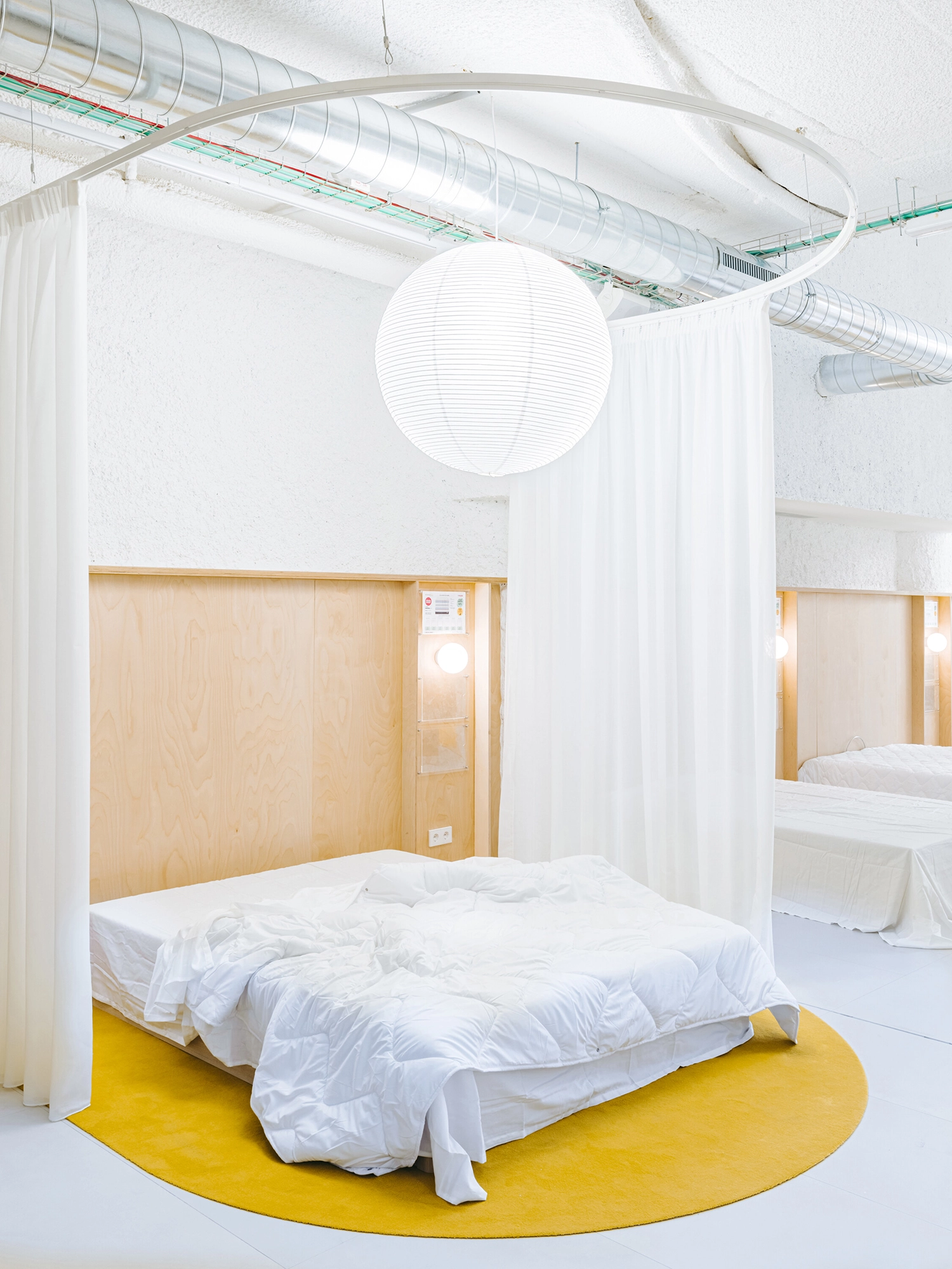 Close-up of a bed setting with a white curtain, yellow rug, and exposed silver ventilation ducts.
