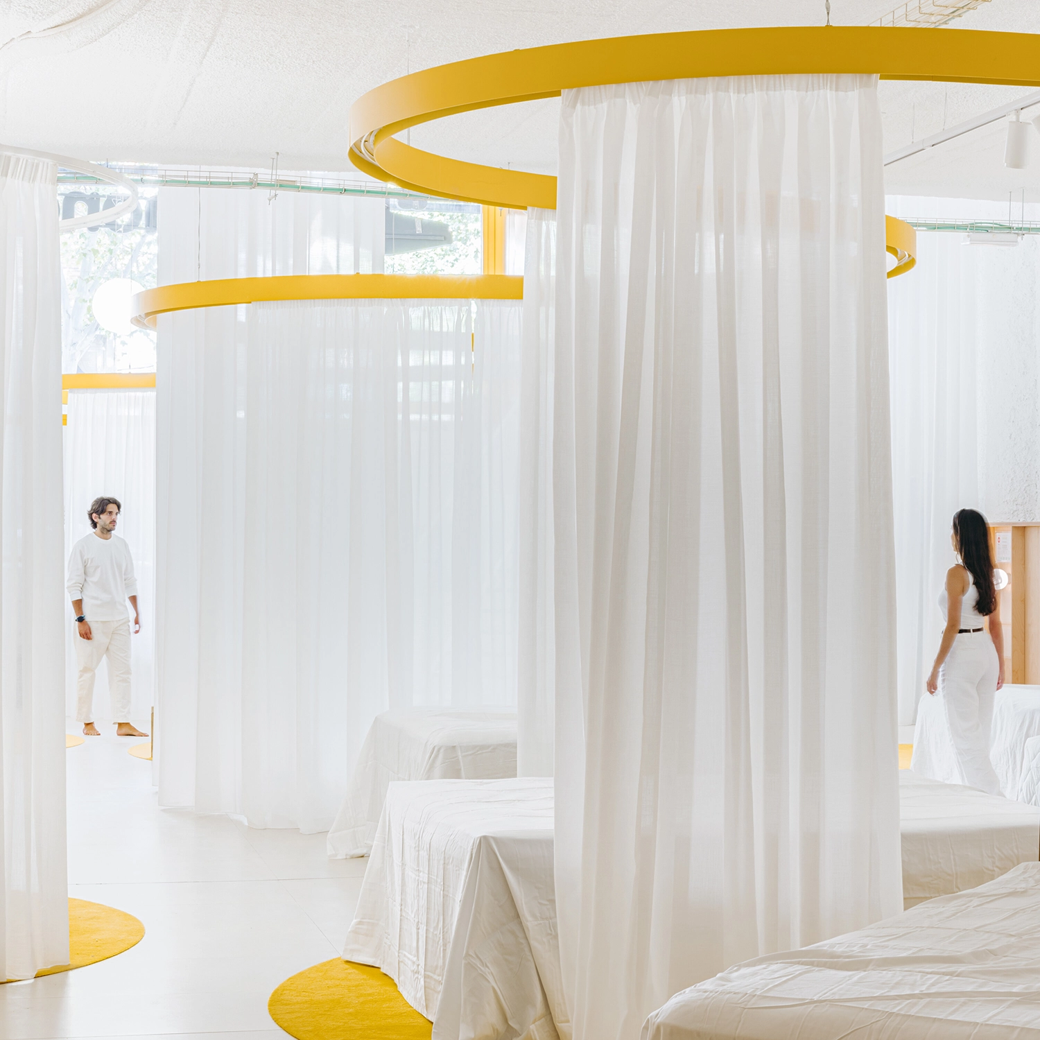 Figures walking through a series of curtain-enclosed mattress displays in Madrid.