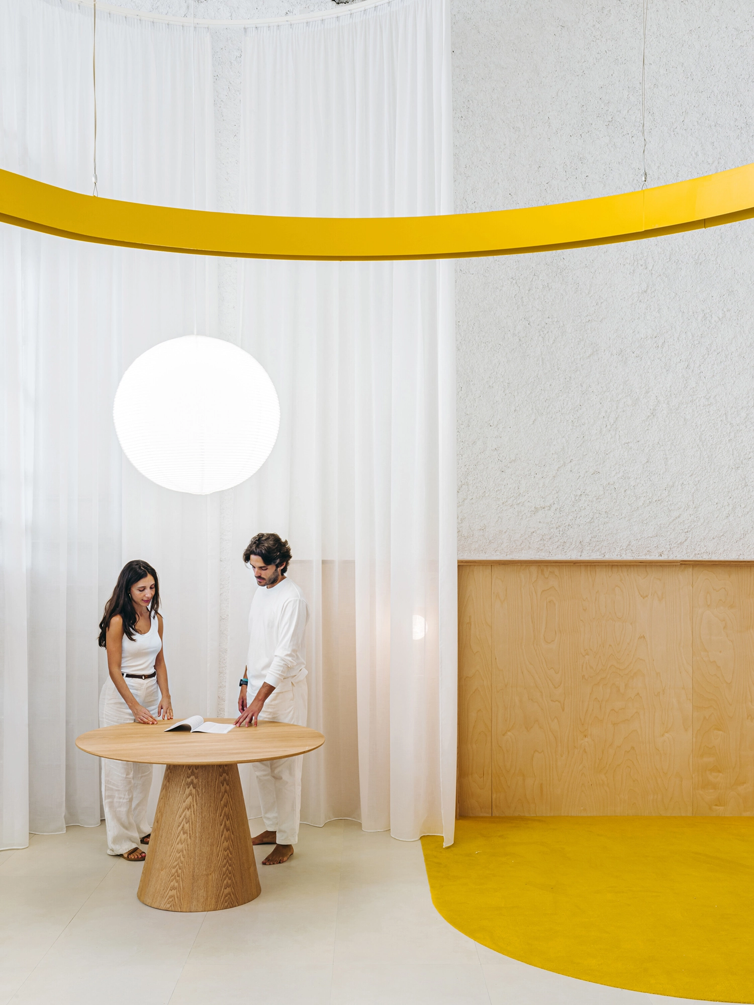 Two people standing at a wooden table in a retail space with a yellow floor and white curtain.