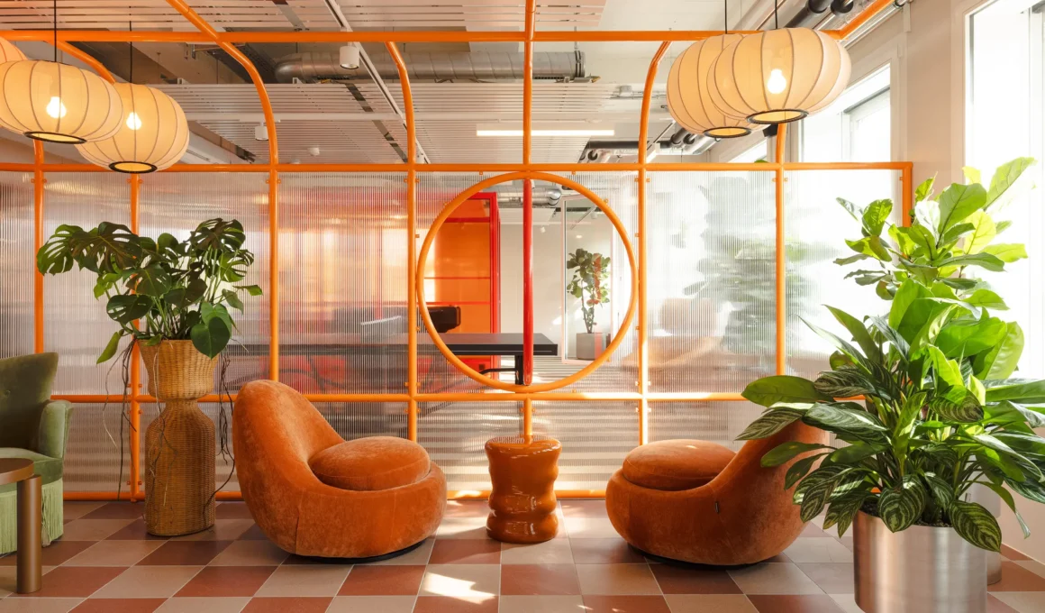 A vibrant workspace interior with striking orange piping frames, ribbed polycarbonate partitions, industrial concrete, plush orange velvet lounge chairs, and a metal planter with a Monstera deliciosa, designed to facilitate casual encounters.