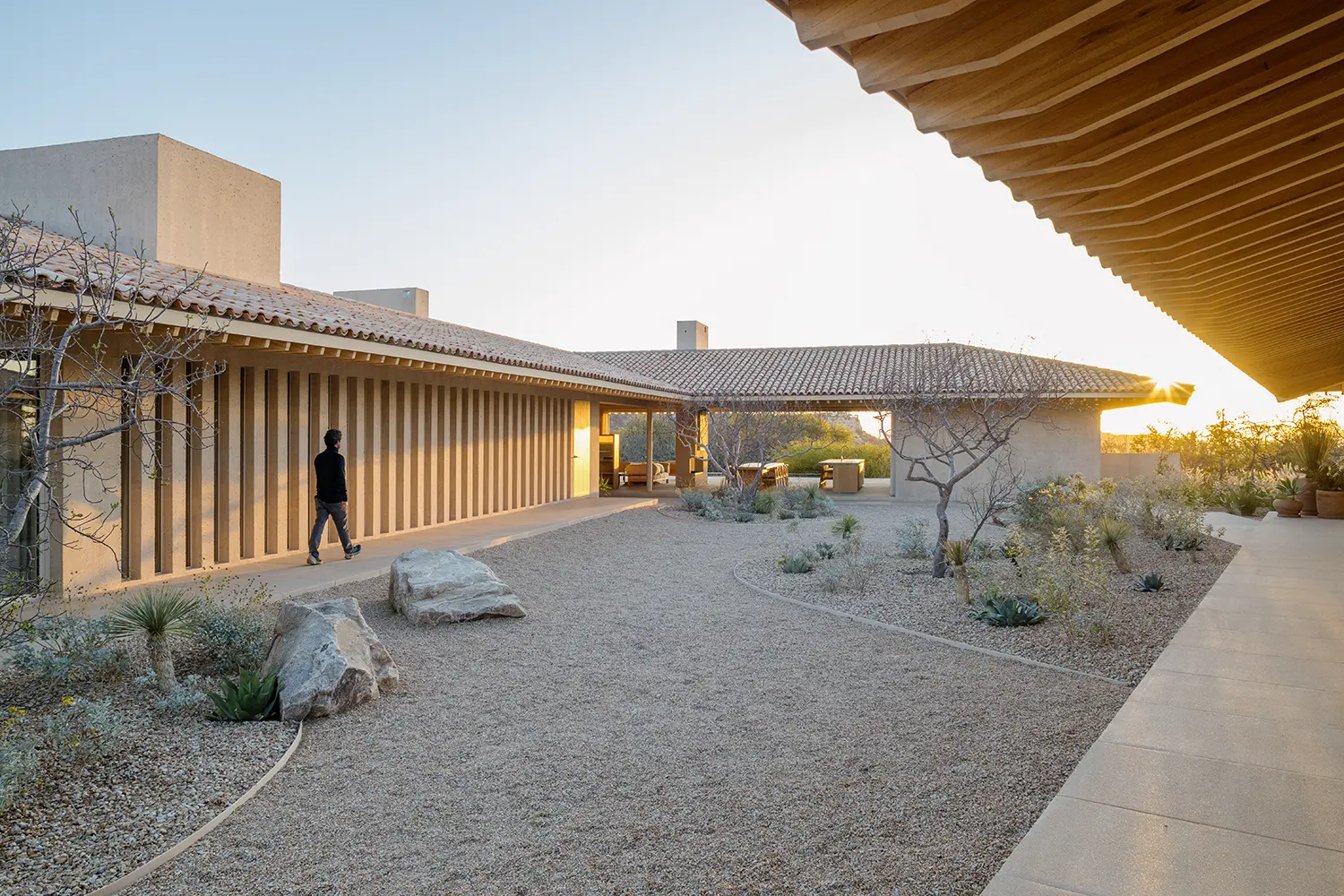 Exterior view of sand-colored concrete volumes and tiled roofs surrounding a central gravel courtyard at sunset.