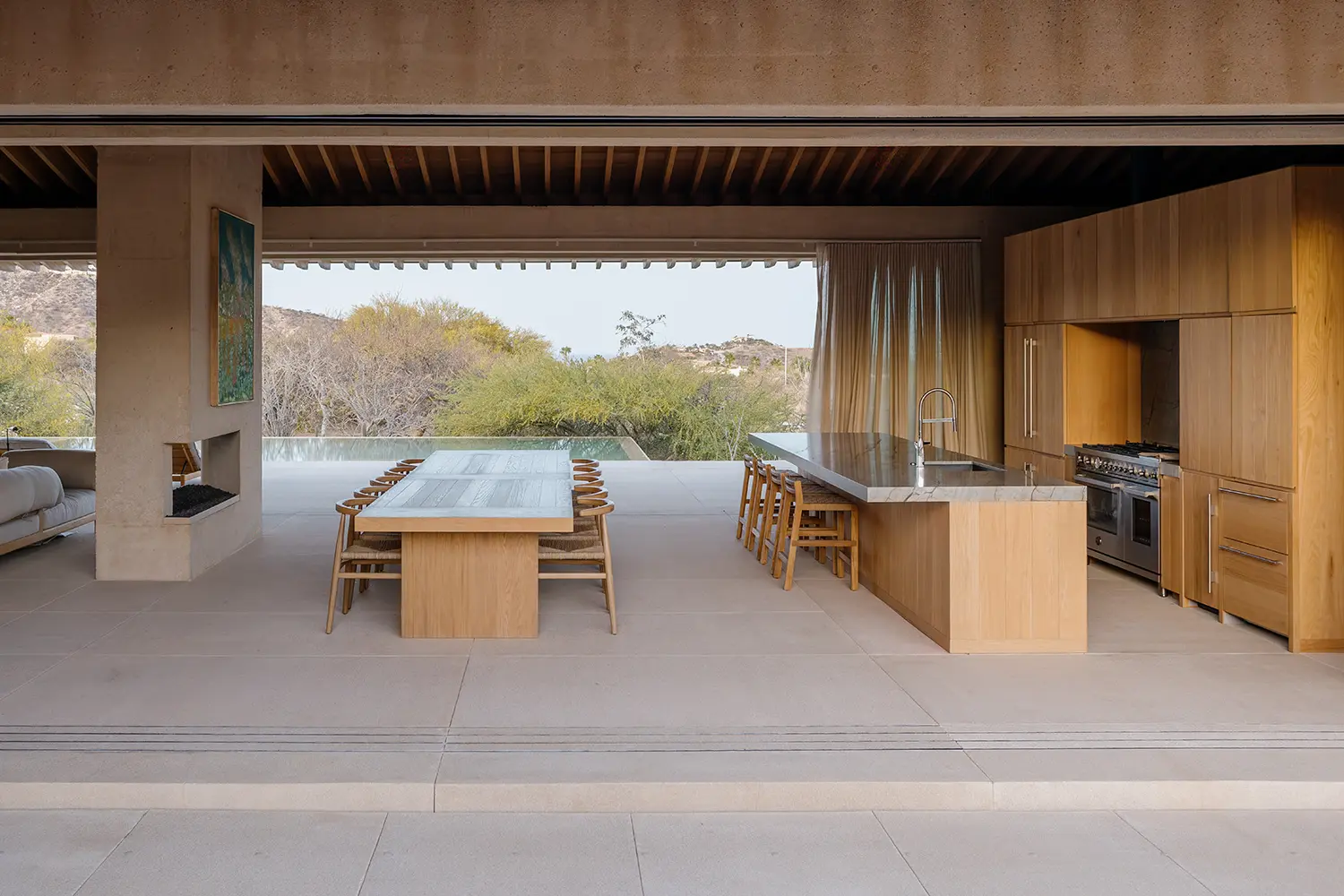 Symmetrical view of a long wooden dining table and kitchen island looking out toward a swimming pool and desert hills.