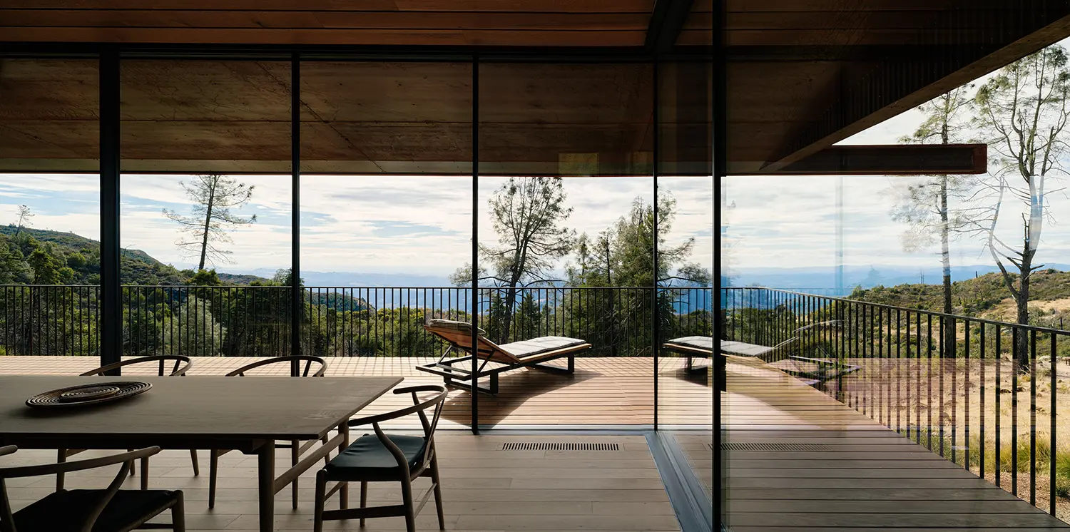 Interior view of a dining room with floor-to-ceiling glass walls overlooking a forested valley.