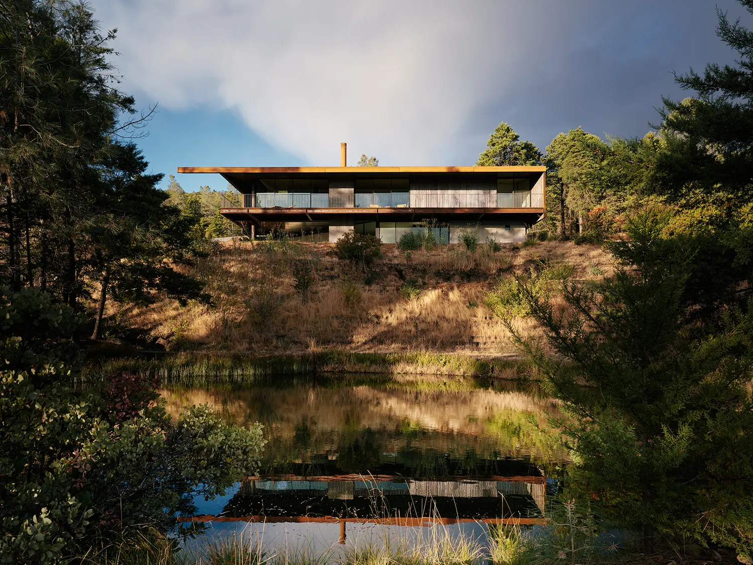 A modern steel and concrete house perched on a hillside above a reflecting pond.