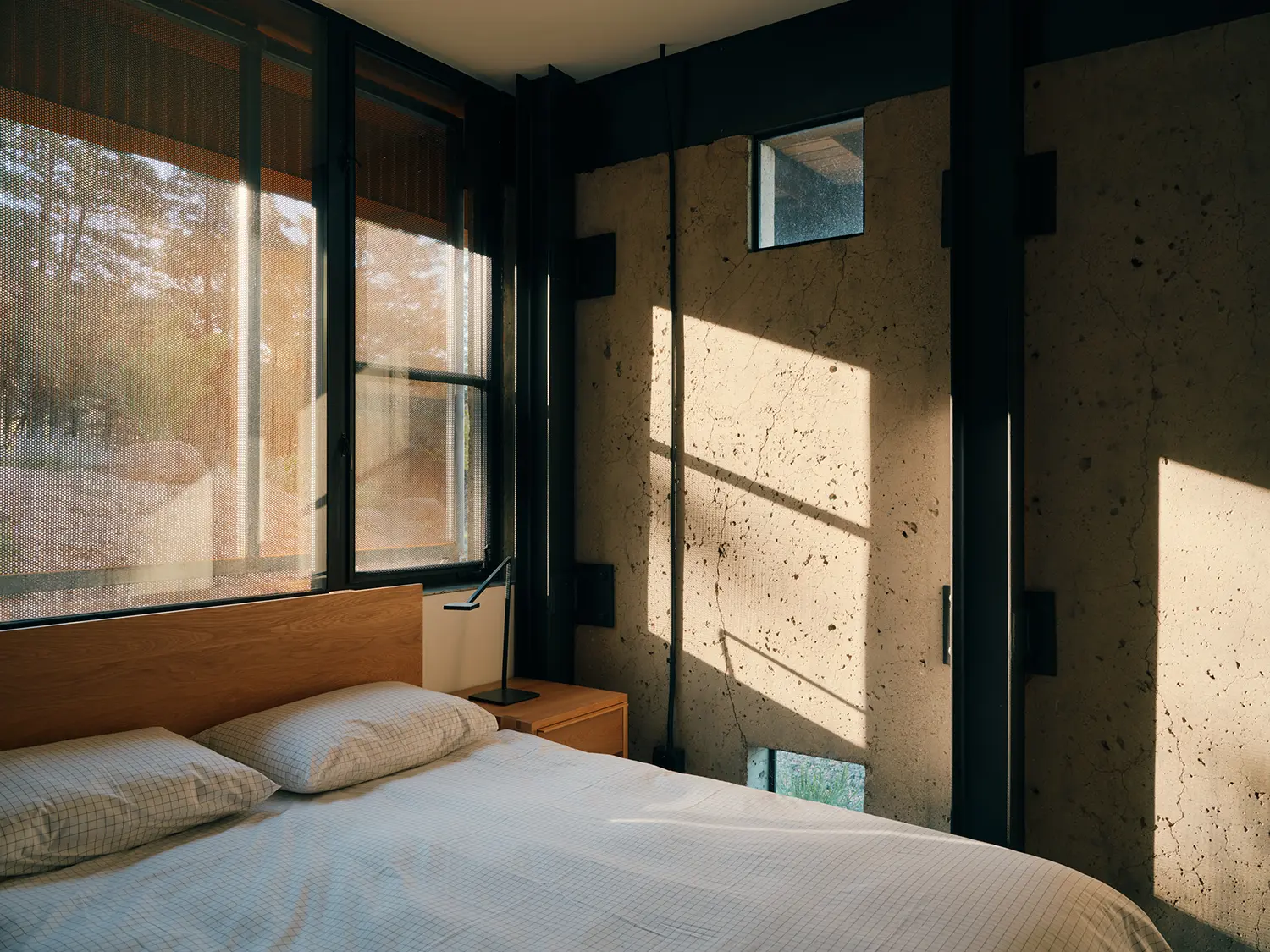 A minimalist bedroom with a raw concrete wall, wooden headboard, and metal screens.