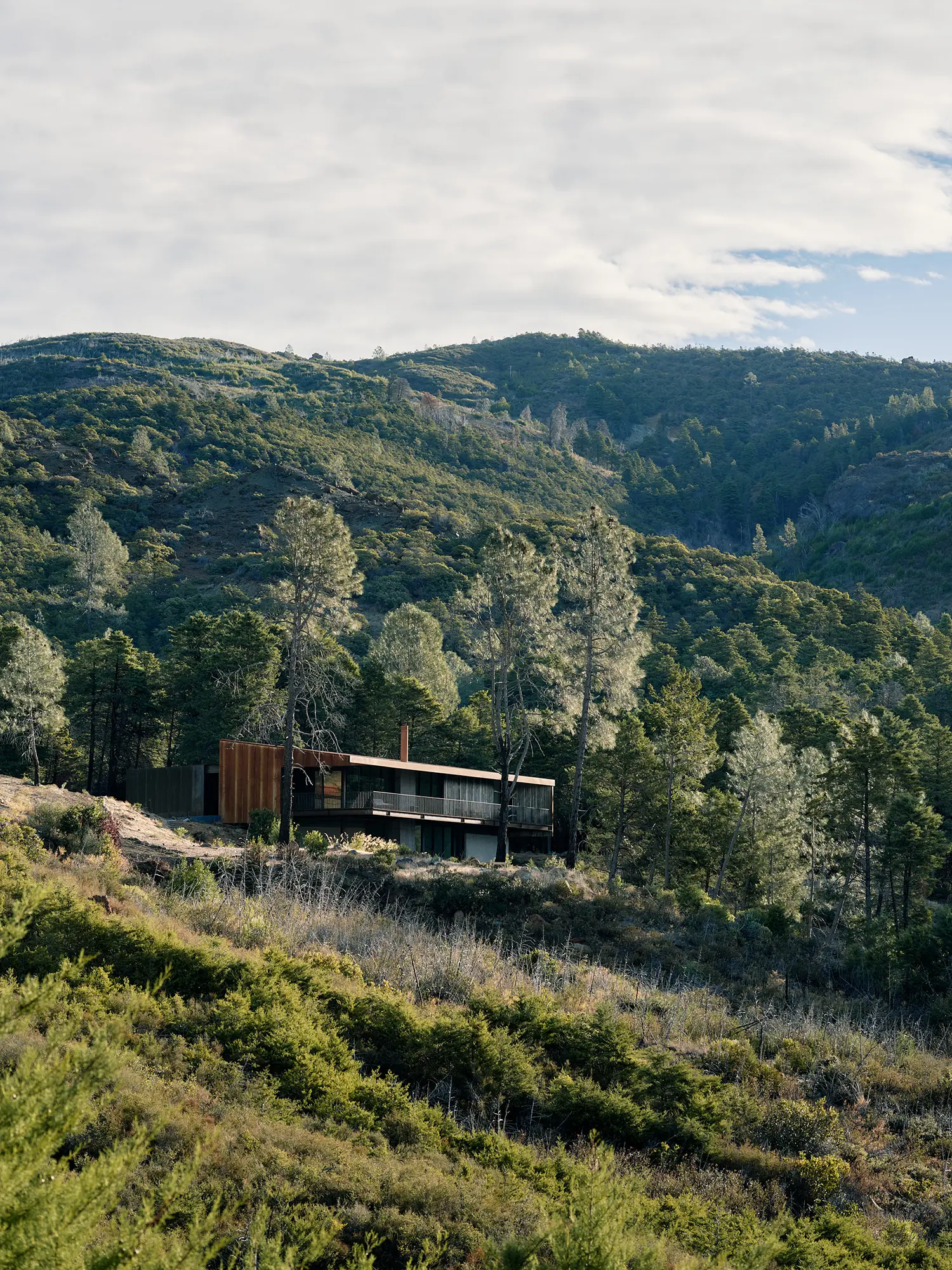 Wide shot of a dark steel house tucked into a dense green mountain landscape.