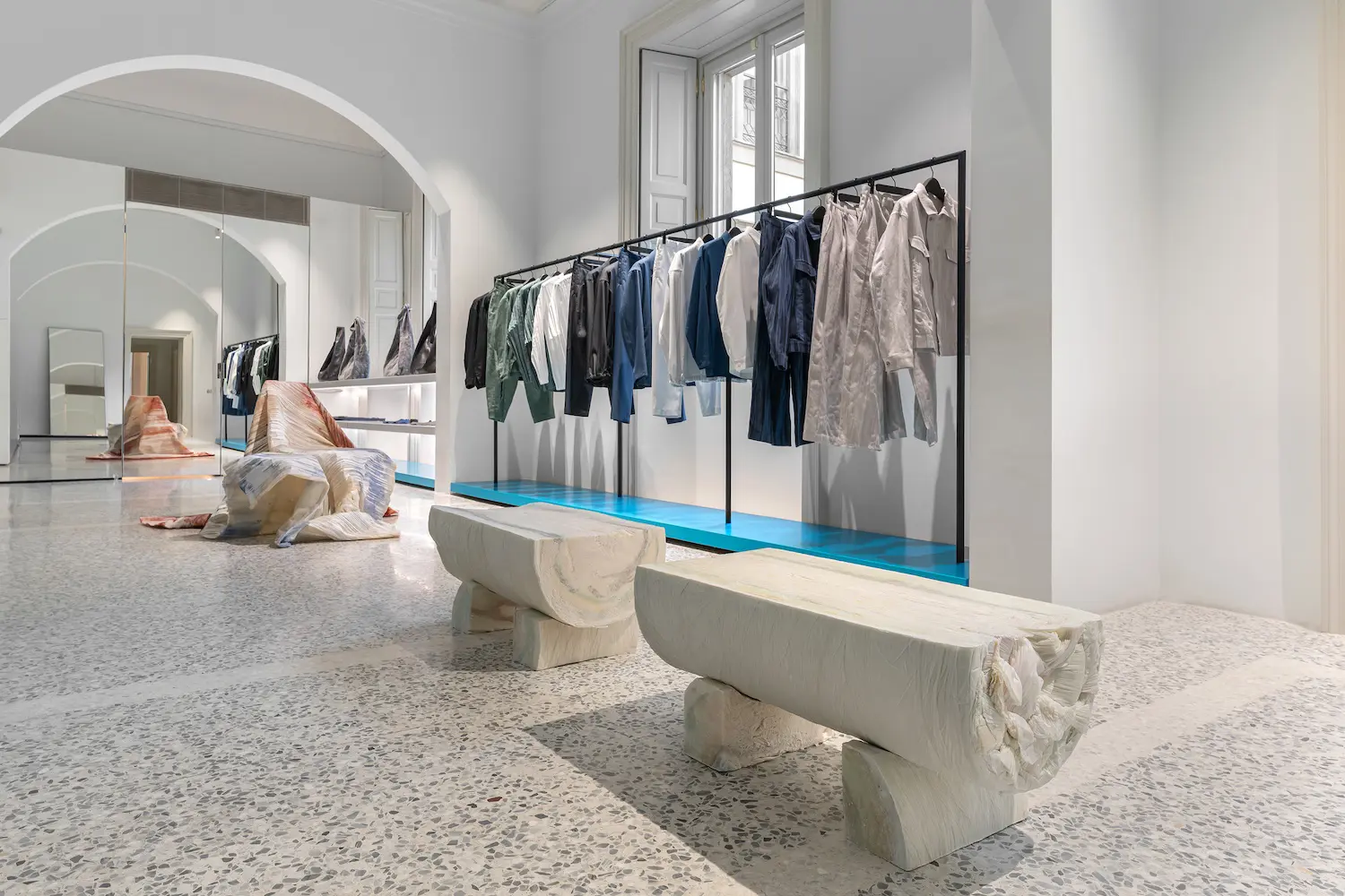 Interior of the ISSEY MIYAKE Milan store featuring long garment racks and two large, solid paper log benches in the foreground.