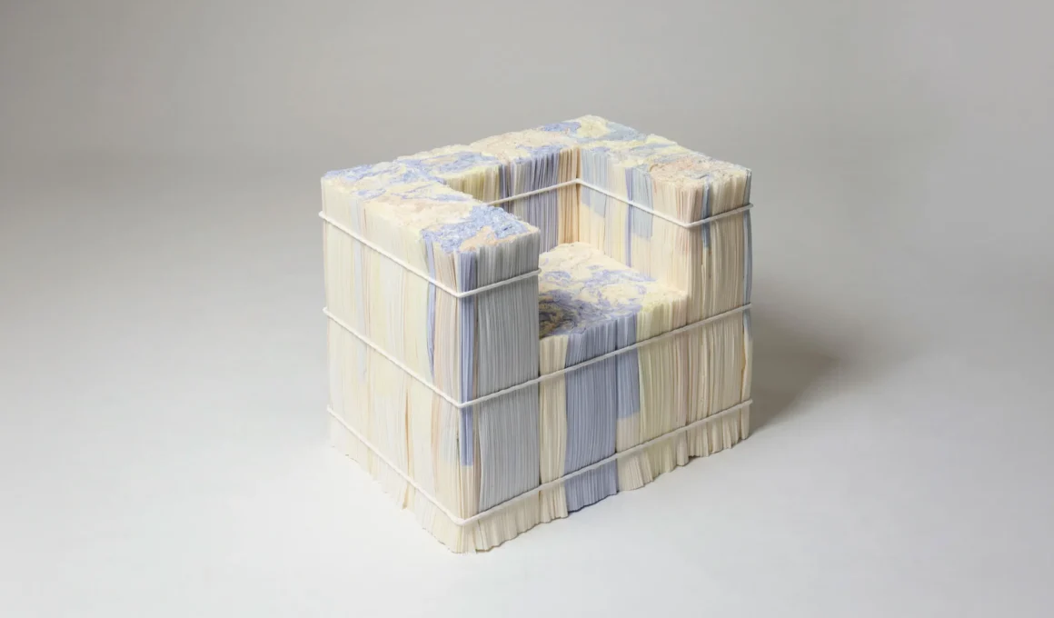 A studio shot of a cubic sculptural armchair constructed from vertical stacks of off-white, soft yellow, and pastel lavender compressed pleated paper. The structure is secured by three horizontal white wire binders and reveals a marbled, textured surface on the top and seat.