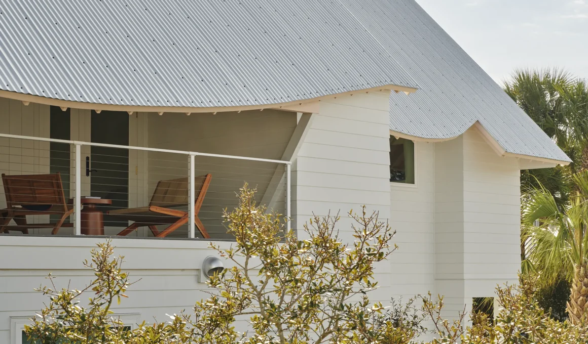 Close-up of a white house with a corrugated metal roof featuring a curved, pink-painted eave and a balcony with wooden lounge chairs.