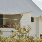 Close-up of a white house with a corrugated metal roof featuring a curved, pink-painted eave and a balcony with wooden lounge chairs.
