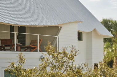 Close-up of a white house with a corrugated metal roof featuring a curved, pink-painted eave and a balcony with wooden lounge chairs.