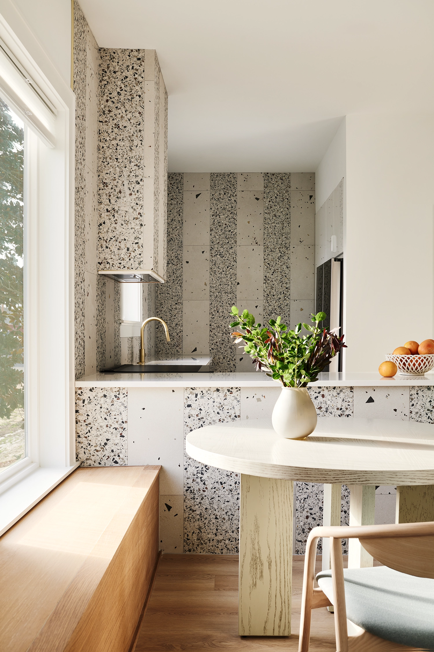 Interior view of a dining nook with a wooden bench, a round table, and a wall featuring vertical strips of speckled terrazzo.
