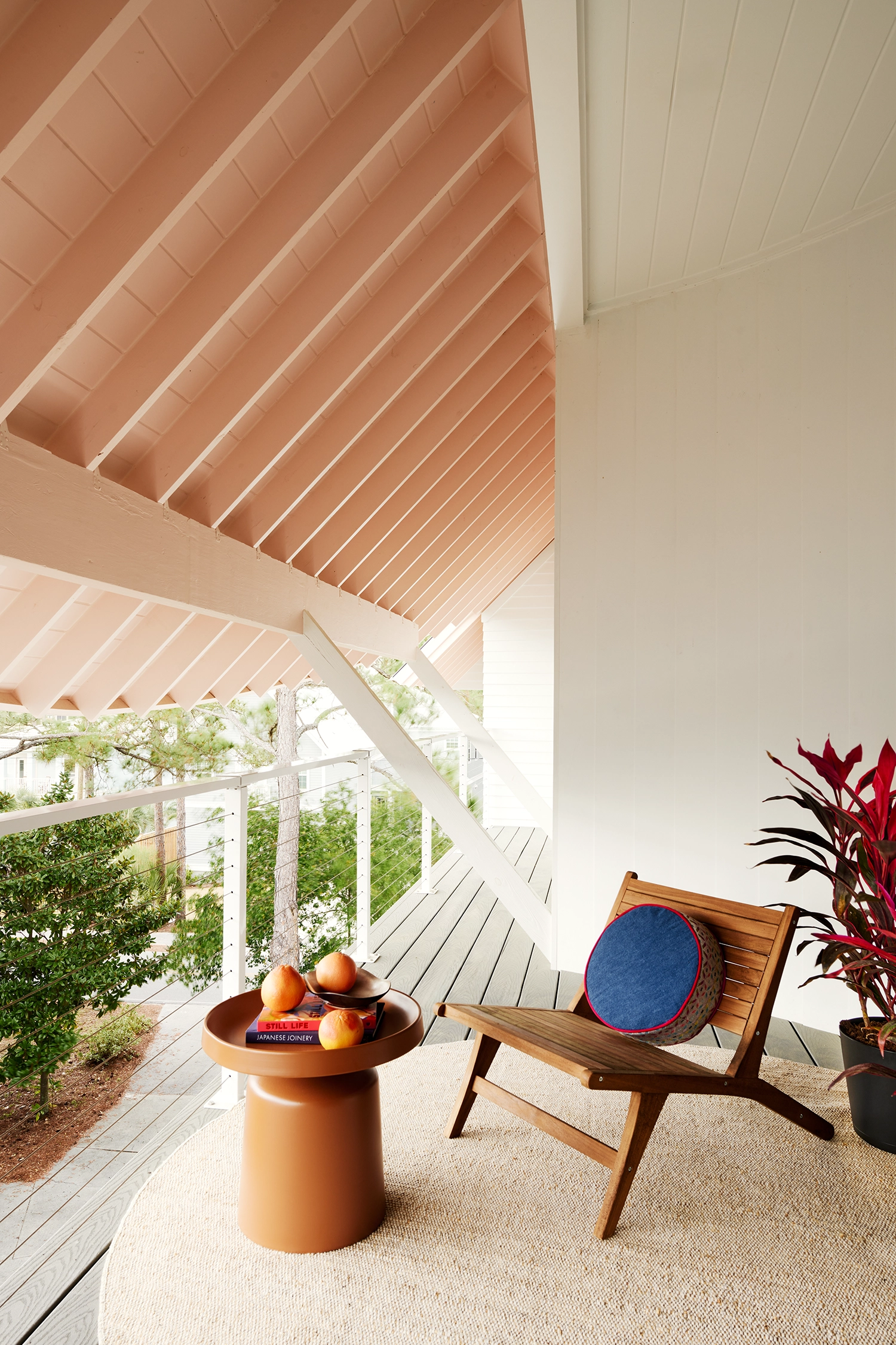 A covered balcony with a pink rafter ceiling, a wooden chair, and a view of surrounding pine trees.