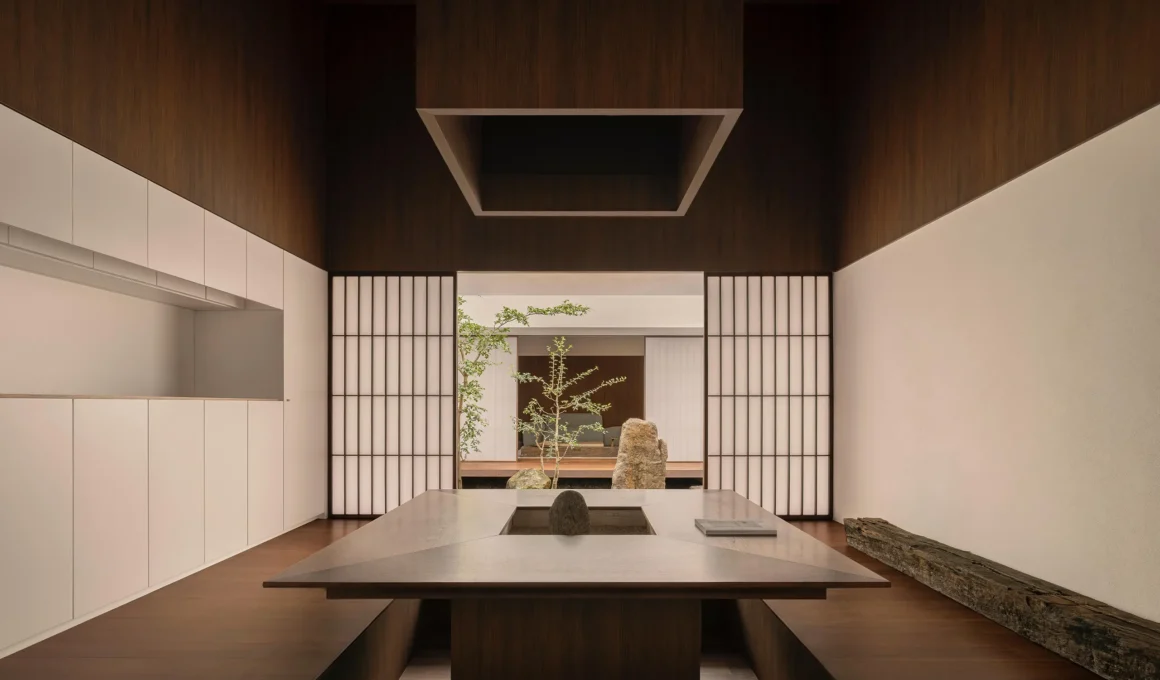 Interior view of a minimalist dining area with a sunken wooden table and shōji screens looking into a courtyard.
