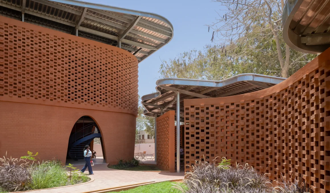 Exterior view of the Goethe-Institut Dakar, showing the curved rammed earth brick facade with perforated lattice patterns. A pedestrian walks on a paved path in the foreground near lush greenery and ornamental purple reeds. In the background, a spiral staircase and a large, metallic wavy canopy roof are visible.