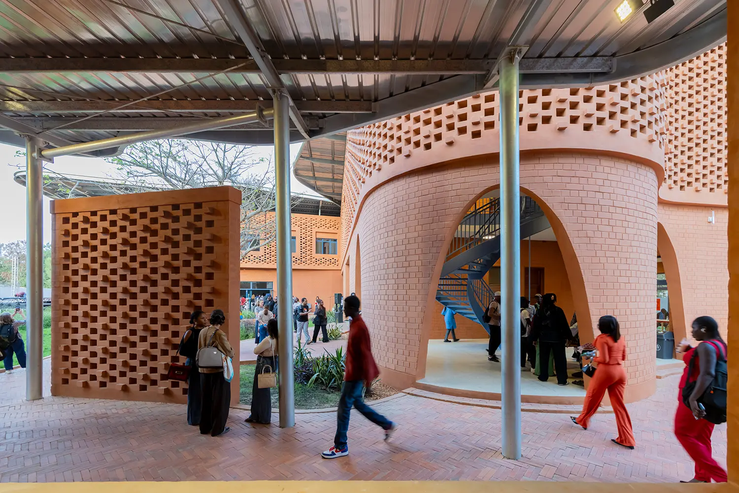 An internal courtyard view, looking out from under the metallic canopy. People are gathered, socializing and walking, near the earth-brick walls and columns. The ground is paved with reddish bricks, and an external spiral staircase is visible.