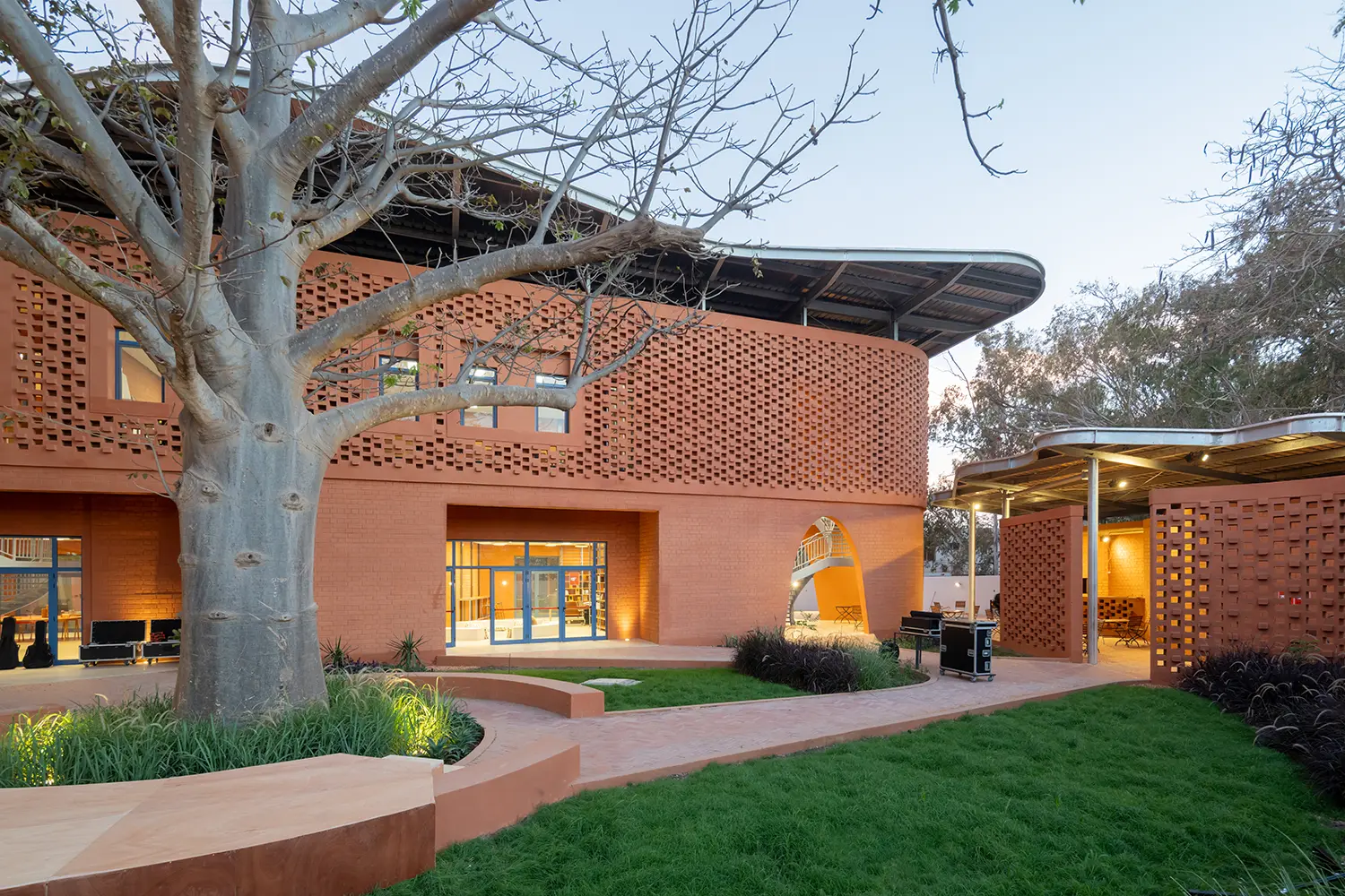 Exterior view of the courtyard at dusk, focusing on a large baobab tree integrated into the design. The earth-brick facade, wavy metal canopy, and garden lighting are all visible, with equipment cases positioned on the path