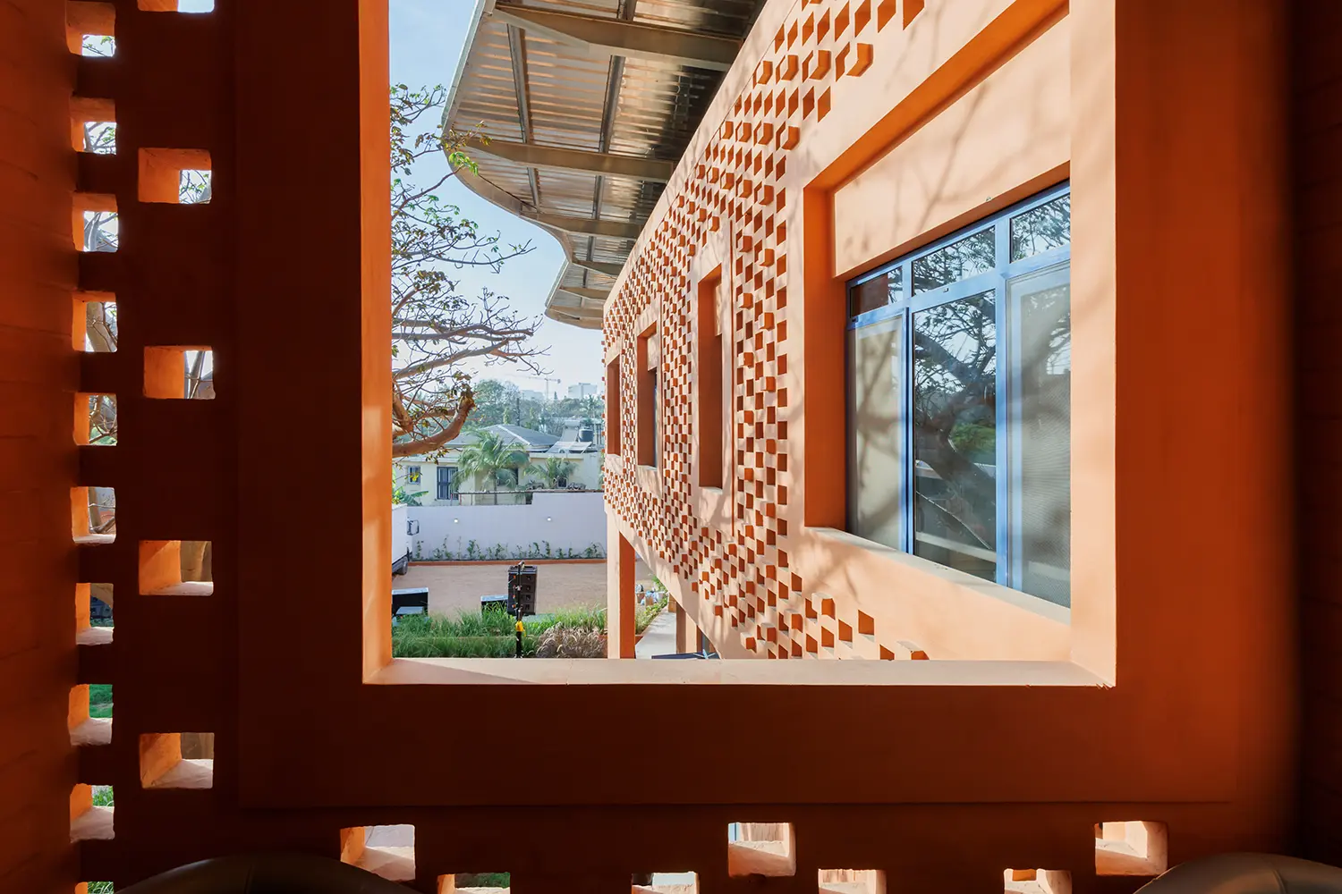 Interior view looking through a large rectangular arched window with perforated brick screens. Beyond the frame, another wing of the earth-brick building and trees are visible under a clear sky.