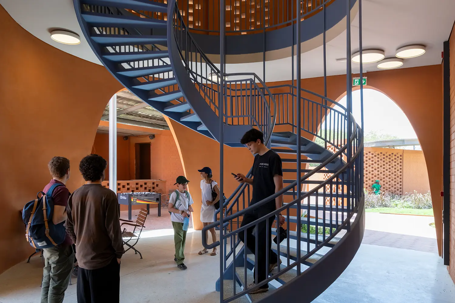 Interior view looking toward an external courtyard, featuring a central black spiral staircase. People stand nearby, and game tables are visible outside under the canopy through an arched opening.