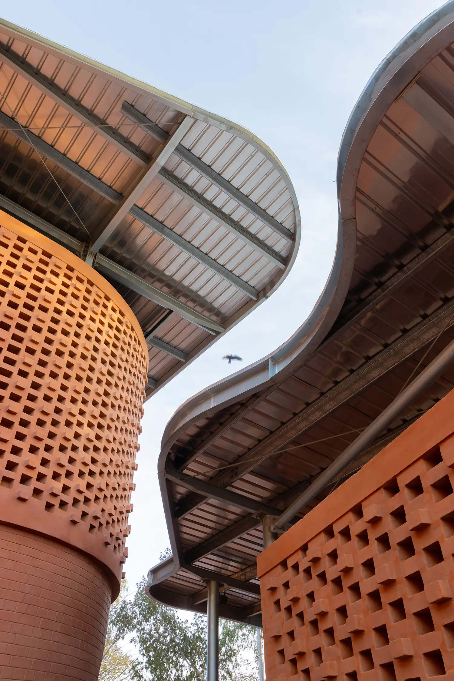 Upward-angle shot of the layered, wavy, corrugated metal canopy roof structure, contrasting with the geometric, perforated lattice brick wall.