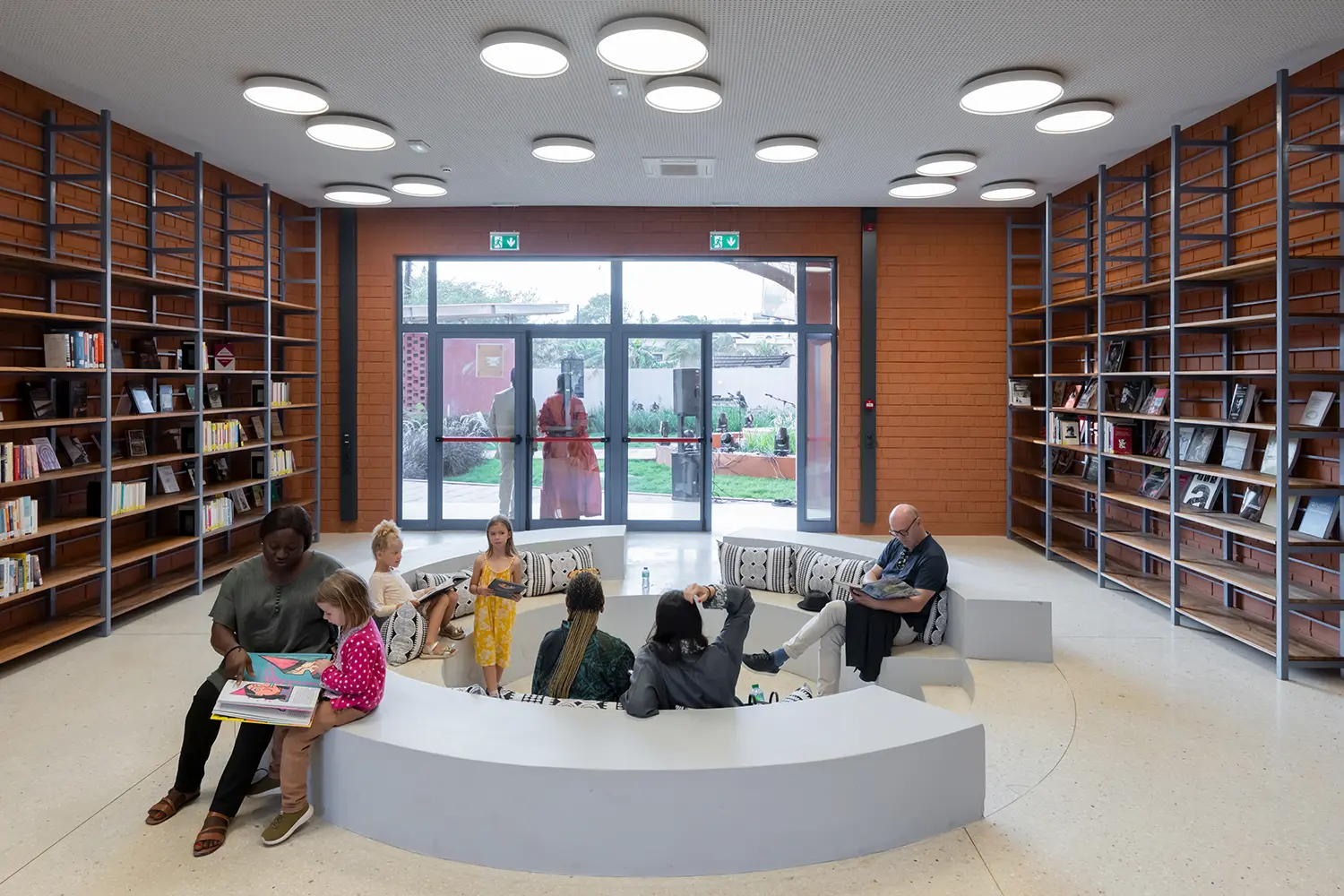 Interior view of a circular library room where a group sits and reads on a central curved bench. The walls are red brick, and the high ceilings feature round flush lights. Bookshelves line the background.