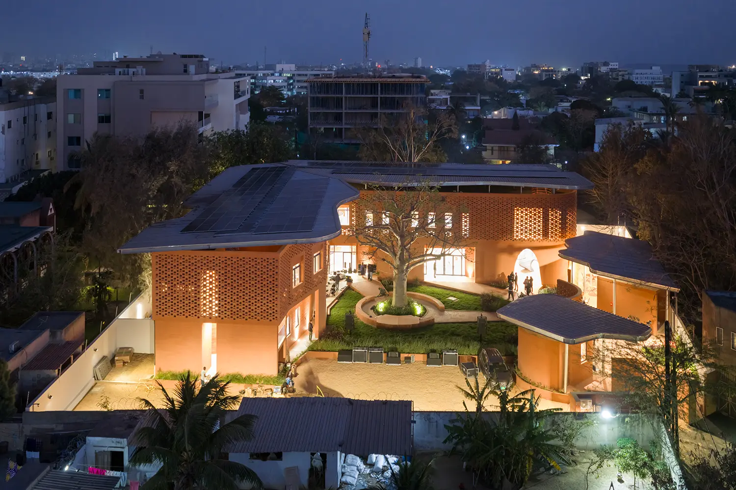 Aerial night photograph of the entire Goethe-Institut complex. The orange earth buildings encircle a central courtyard with a large baobab tree. Solar panels cover the metal canopy roofs.