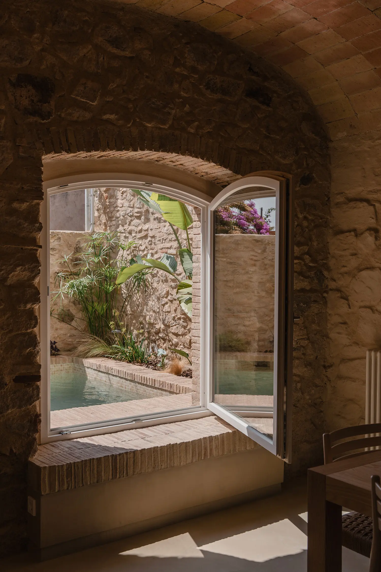 A view through an arched white window looking out toward a small turquoise plunge pool and stone walls.