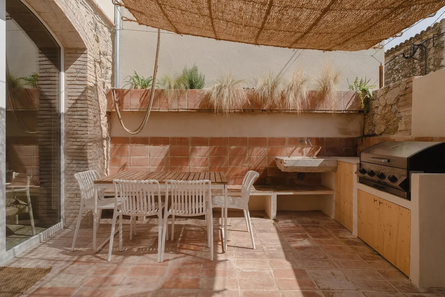 An outdoor patio with a wooden table, white chairs, and a built-in grill under a burlap canopy.