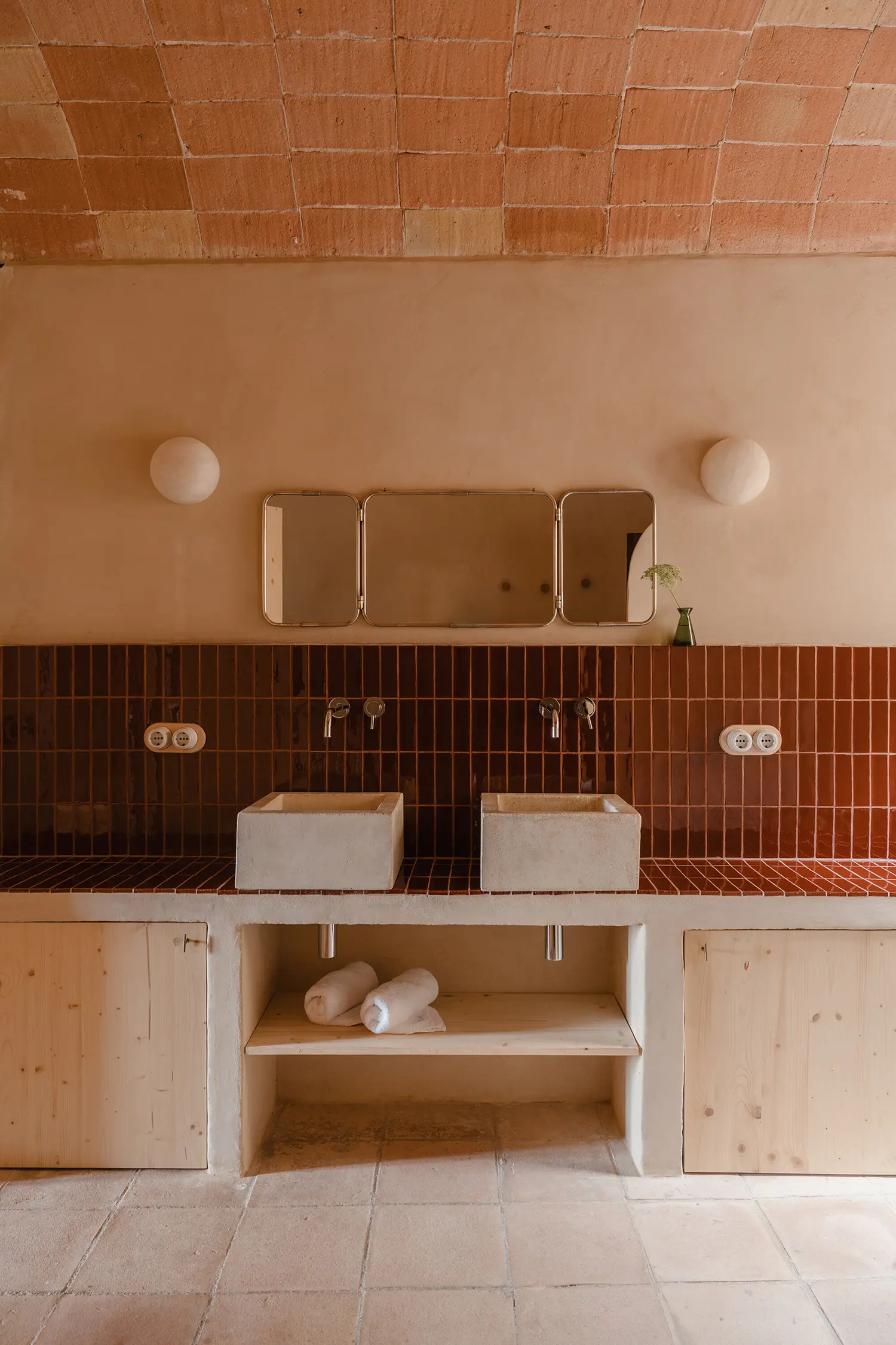 A bathroom with two stone vessel sinks, deep red tiles, and a triptych mirror.