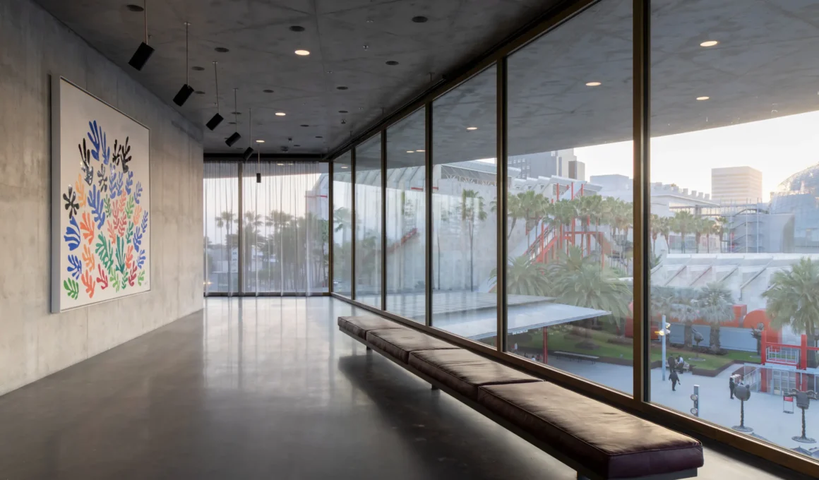 Wide-angle view of a concrete gallery interior with floor-to-ceiling windows and a colorful abstract painting on a raw concrete wall.