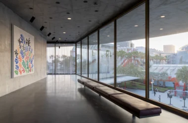 Wide-angle view of a concrete gallery interior with floor-to-ceiling windows and a colorful abstract painting on a raw concrete wall.