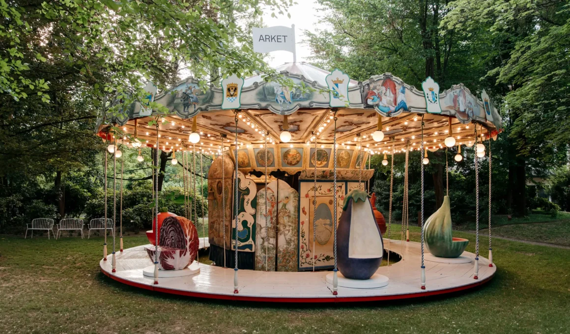 Wide shot of the Laila Gohar x ARKET antique carousel with oversized vegetable seats in a green park.