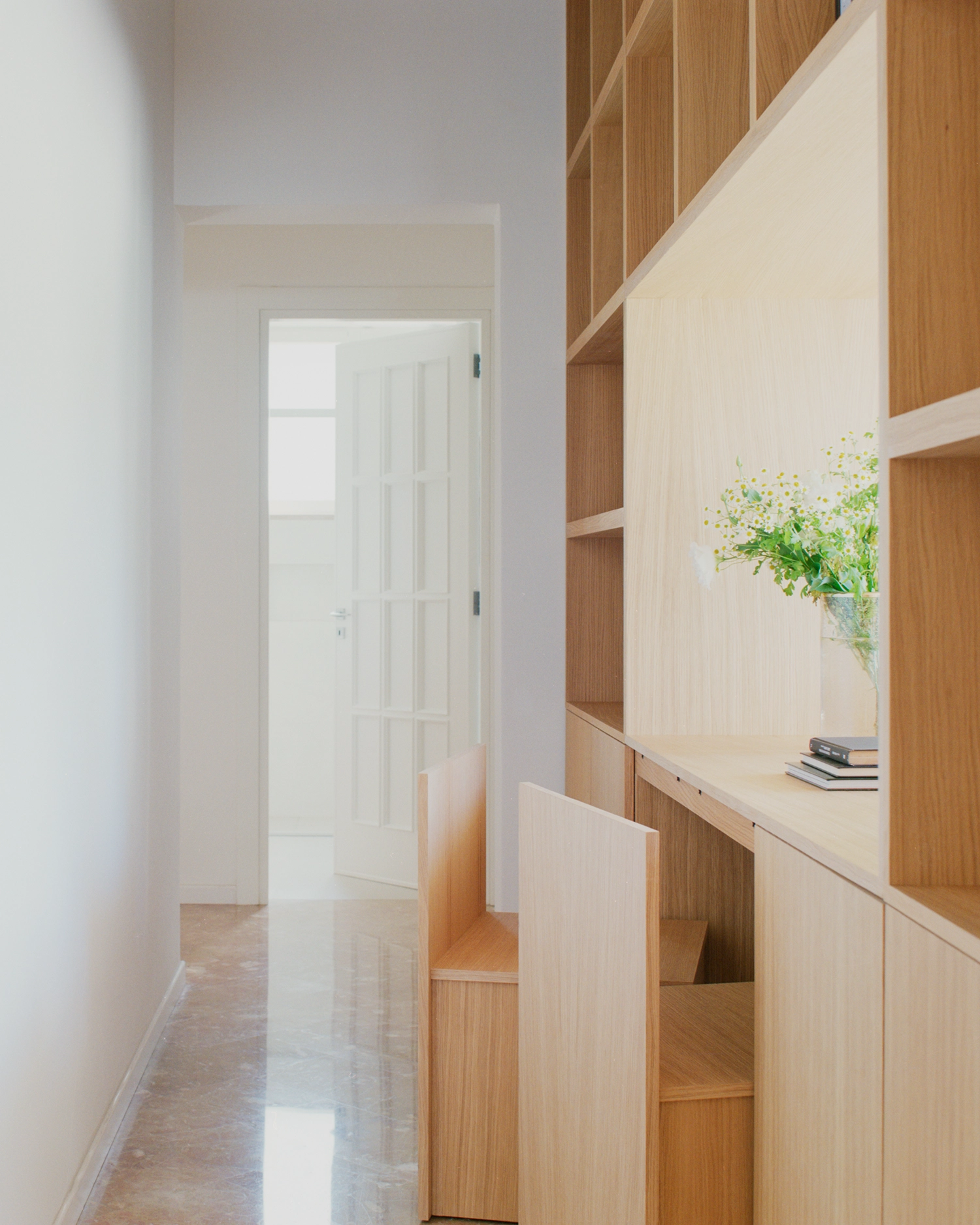 Perspective of a white corridor in Lèvanzo house with built-in oak shelving and a lit internal window.