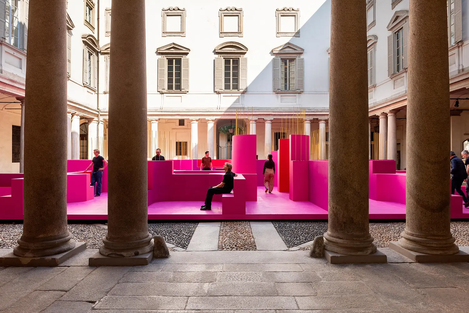Eye-level view through courtyard columns of the pink installation with visitors sitting on integrated benches.