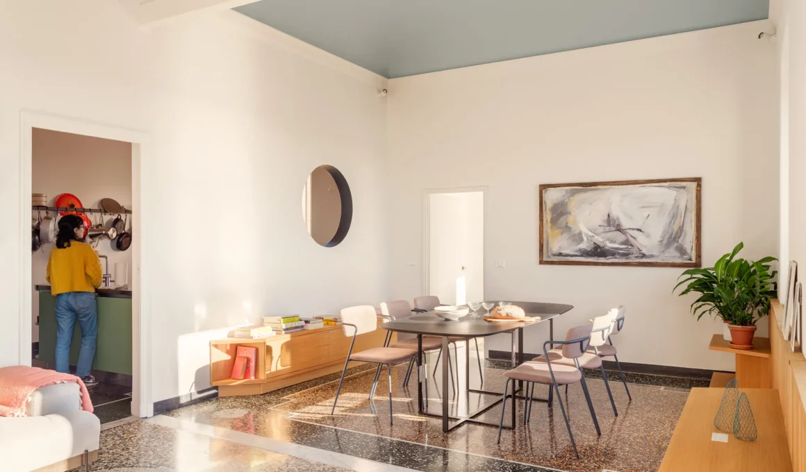 A sunlit dining room with a pale blue ceiling, terrazzo flooring, and a round porthole window looking into the kitchen.