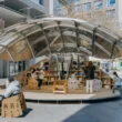 A wide shot of the Shell Book Pavilion by LUO studio fully open in a sunlit plaza, showing children and adults sitting on modular chairs under a large metallic canopy.