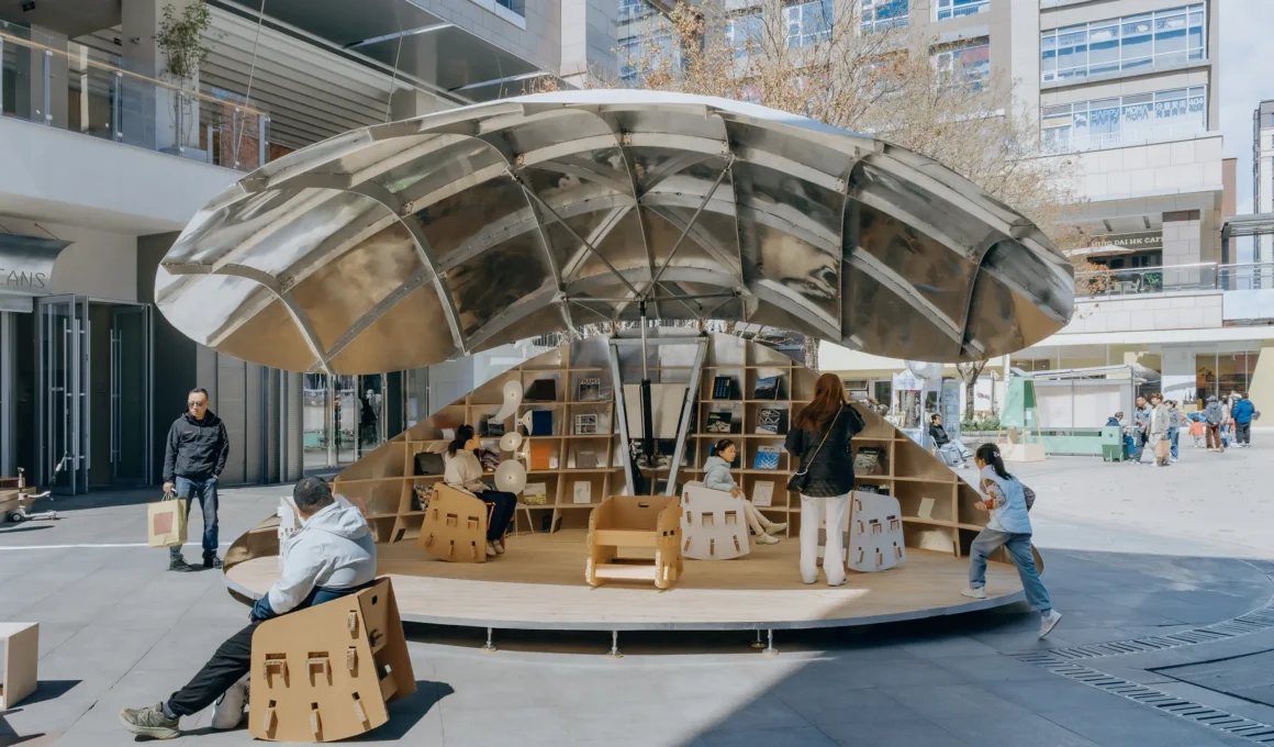 A wide shot of the Shell Book Pavilion by LUO studio fully open in a sunlit plaza, showing children and adults sitting on modular chairs under a large metallic canopy.