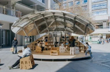 A wide shot of the Shell Book Pavilion by LUO studio fully open in a sunlit plaza, showing children and adults sitting on modular chairs under a large metallic canopy.