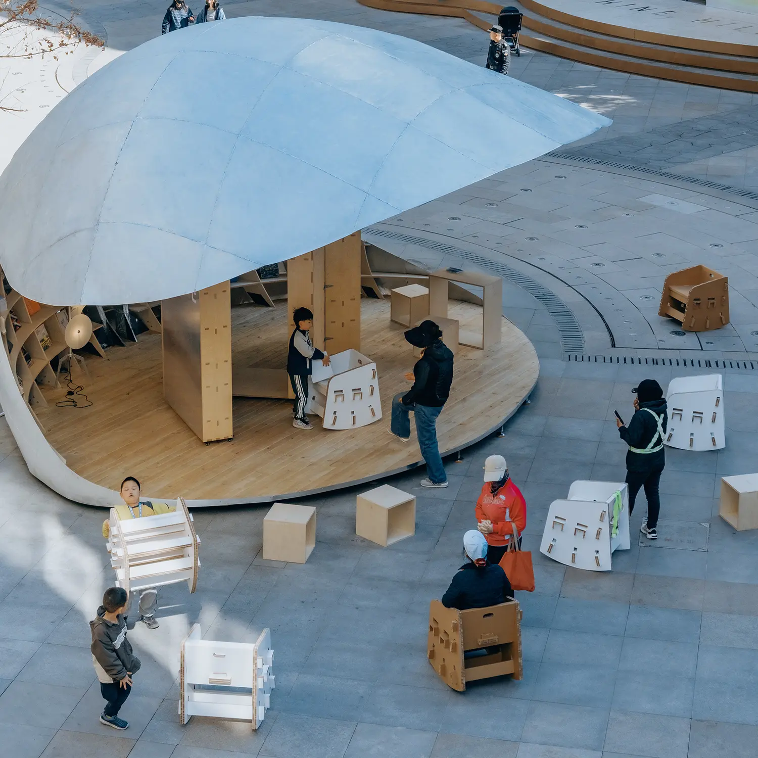 An overhead view of the Shell Book Pavilion in a public square, with children playing with white and brown modular cardboard-style chairs around a circular wooden base.