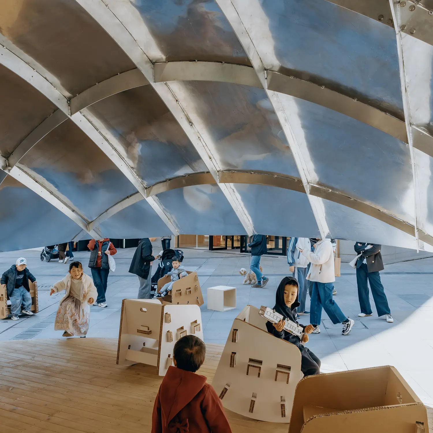 A low-angle shot from inside the pavilion looking out, showing the ribbed metallic structure of the roof and children playing on the wooden deck.