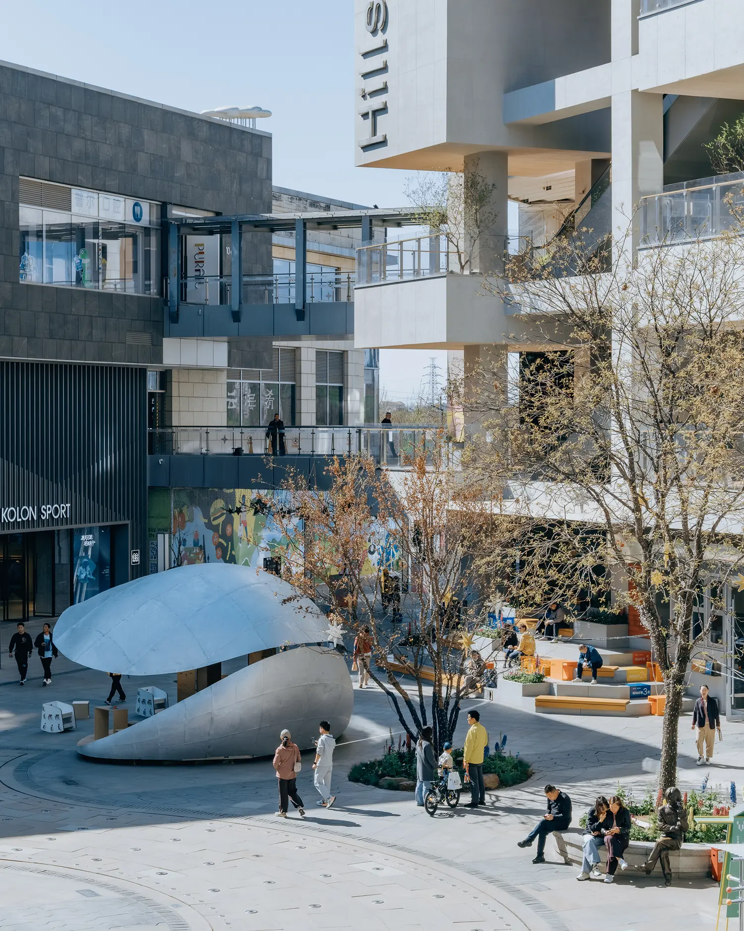 An elevated view of the Shine Hills plaza in Beijing, featuring the silver Shell Book Pavilion nestled among modern commercial buildings and trees.