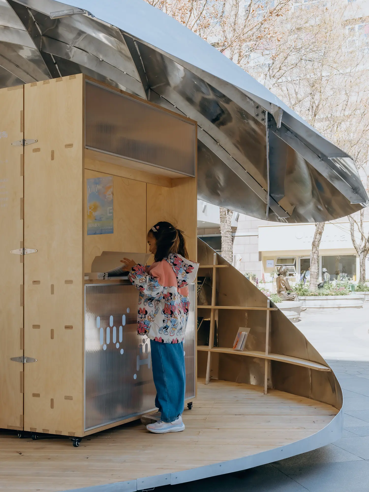 A close-up of a child reading a book inside the wooden interior of the Shell Book Pavilion, with sunlight hitting the metallic roof in the background.