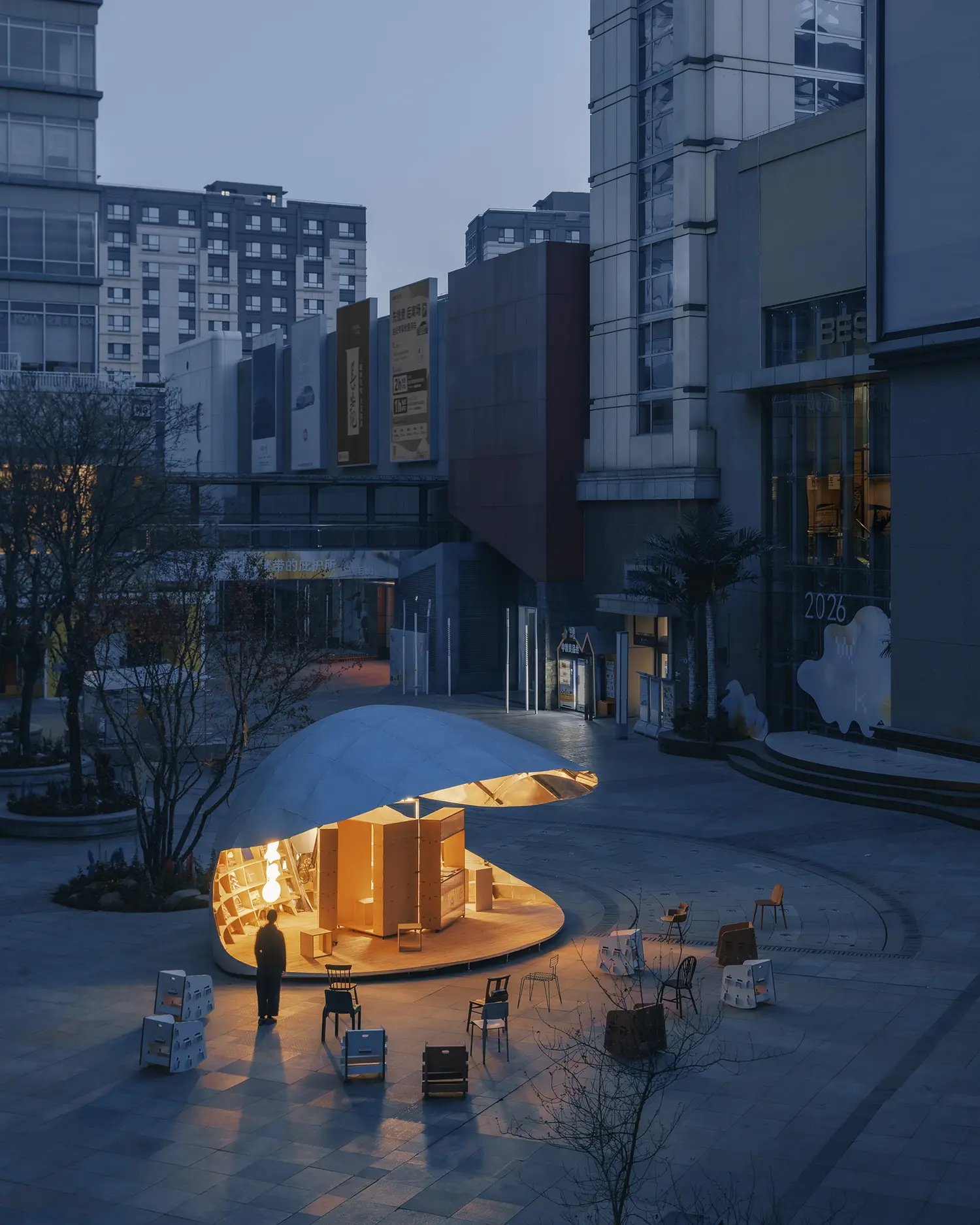 A twilight shot of the Shell Book Pavilion illuminated from within, casting a warm yellow glow across the dark plaza with scattered chairs in the foreground.
