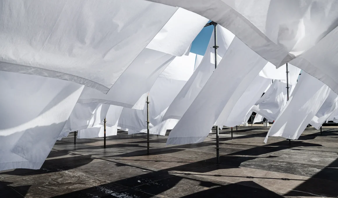 Close-up of white fabric sheets billowing in the wind against a blue sky, supported by metal scaffolding poles.