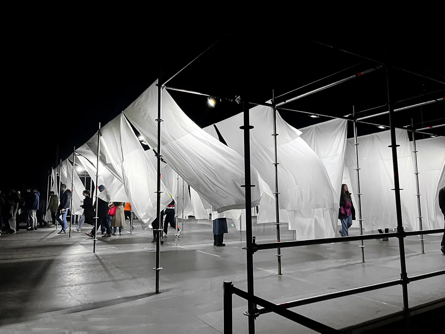 Night view of visitors walking through a maze of white sheets supported by an industrial metal frame.