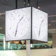Large cubic digital clock suspended from the ceiling in Schiphol Lounge 1, displaying a top-down video of people forming clock hands.