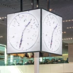 Large cubic digital clock suspended from the ceiling in Schiphol Lounge 1, displaying a top-down video of people forming clock hands.