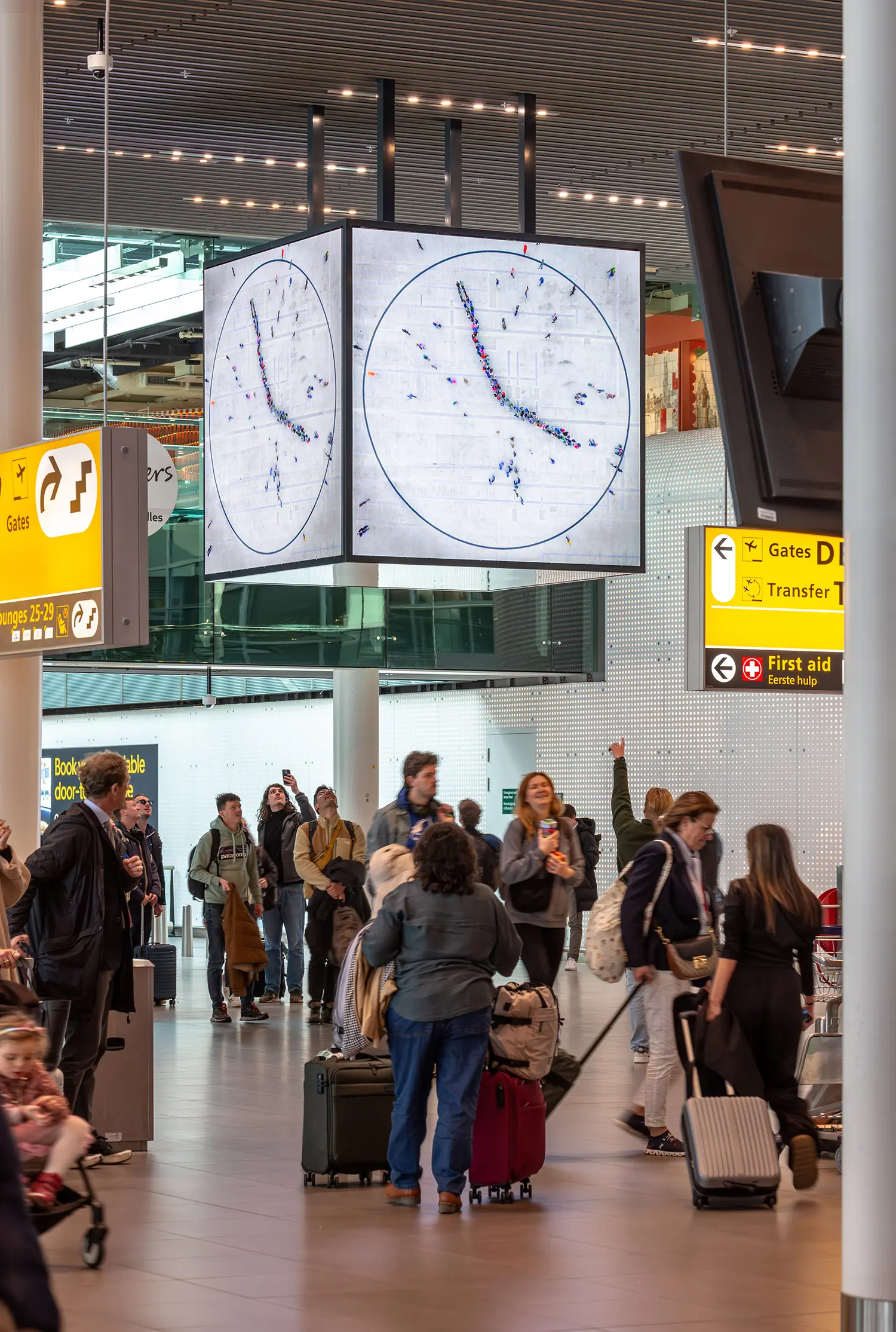 Wide shot of travelers in Lounge 1 looking up at the Maarten Baas People’s Clock, with yellow airport signage in the foreground.
