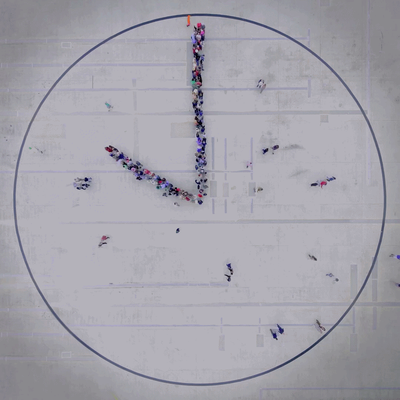Aerial top-down view of Maarten Baas's People's Clock showing hundreds of airport staff in uniform forming two rotating clock hands inside a large blue circle.
