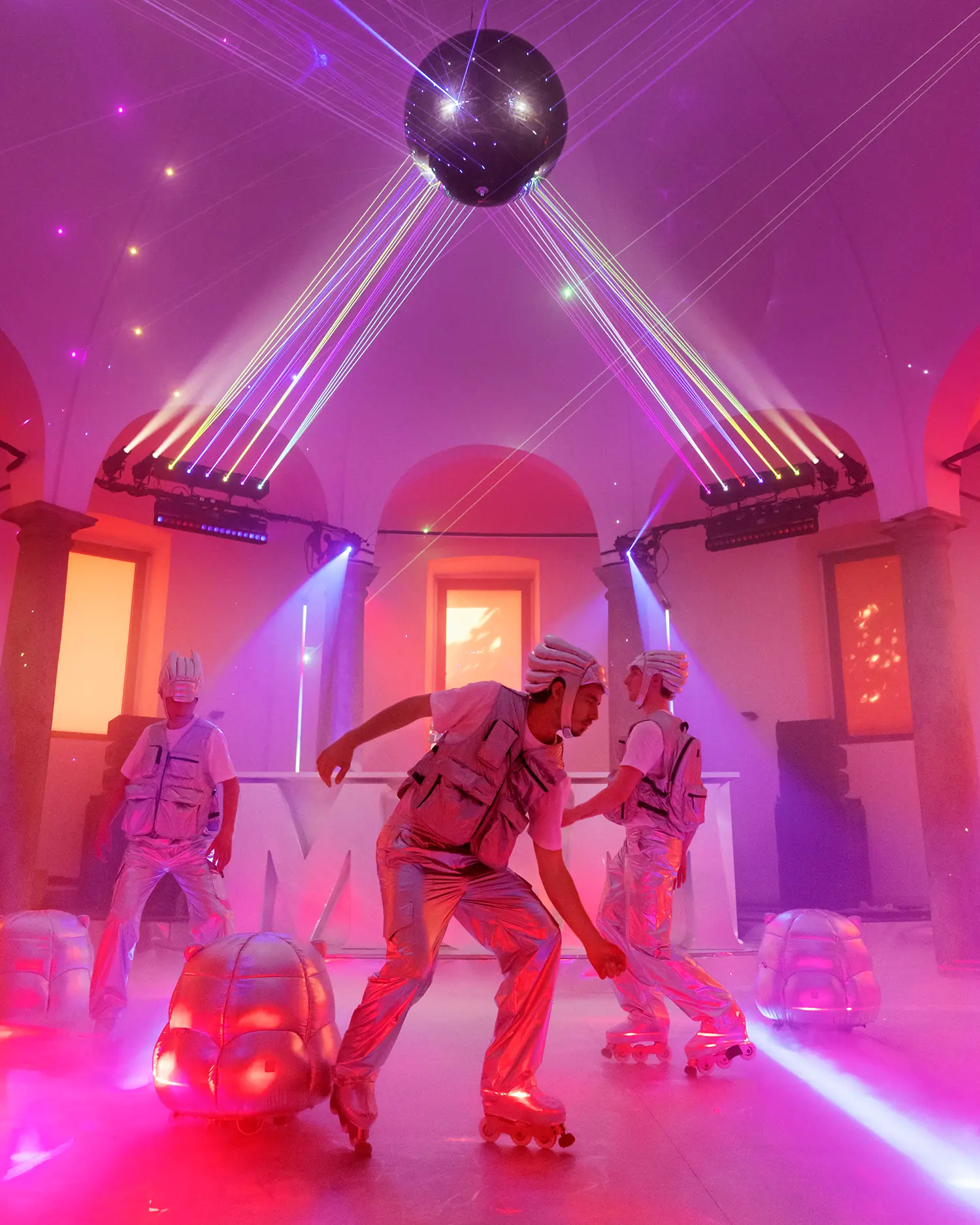Three performers on roller skates in silver outfits dancing under a disco ball and laser lights.